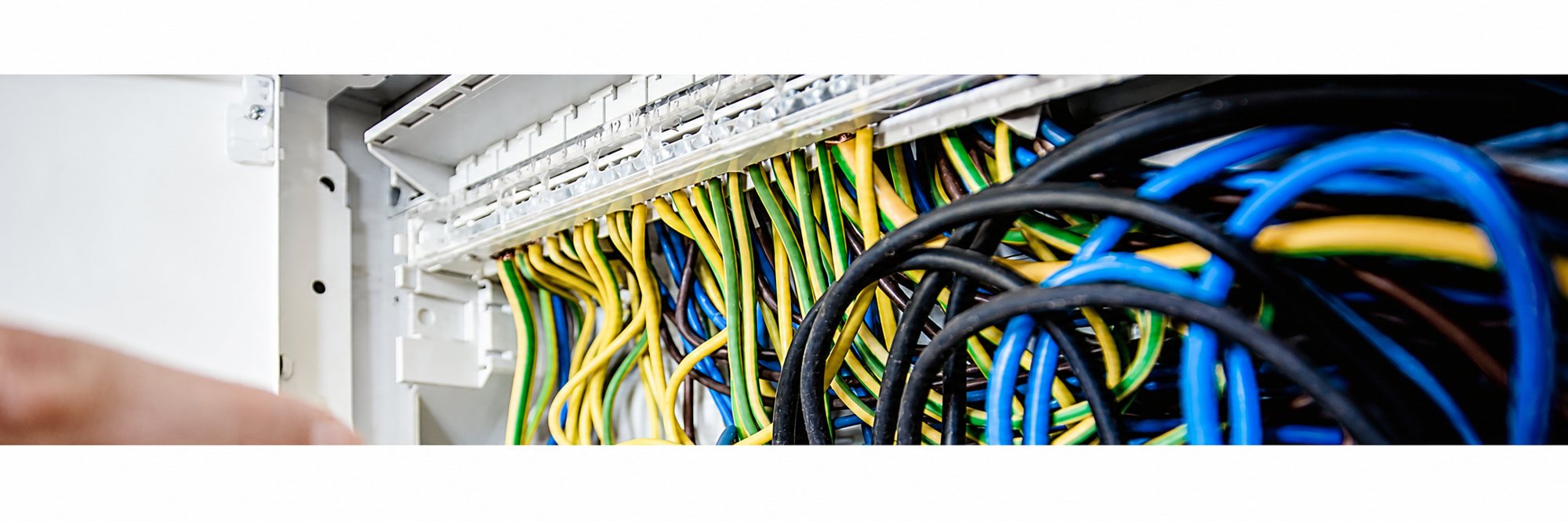 Gros plan sur des fils électriques jaunes, verts, bleus et noirs raccordés dans un tableau électrique blanc, avec un doigt flou au premier plan.