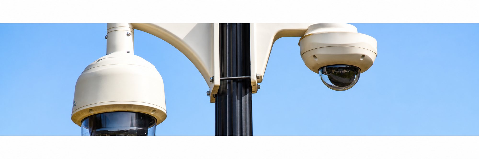 Deux caméras de surveillance de type dôme, de couleur beige, fixées sur un poteau noir devant un ciel bleu dégagé.