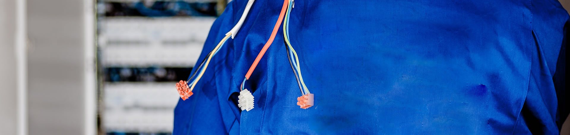 Un électricien en bleu de travail bleu tient plusieurs fils électriques munis de connecteurs orange et blancs devant un tableau électrique flou.
