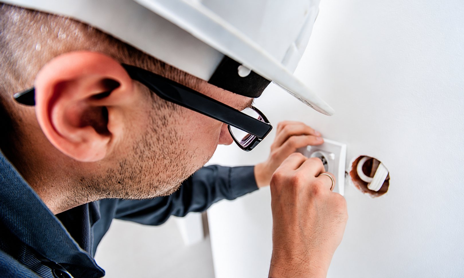 Un électricien portant un casque blanc et des lunettes fixe une prise électrique dans un mur blanc.