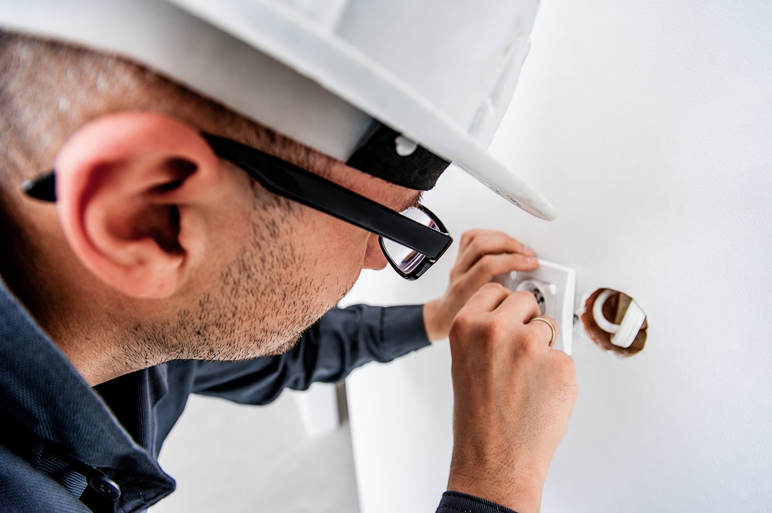 Un électricien portant un casque blanc et des lunettes fixe une prise électrique encastrée dans un mur blanc.