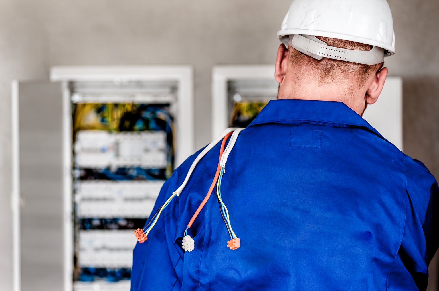 Vu de dos, un électricien en combinaison bleue et casque blanc, avec des fils électriques sur l'épaule, se tient devant un tableau électrique ouvert.