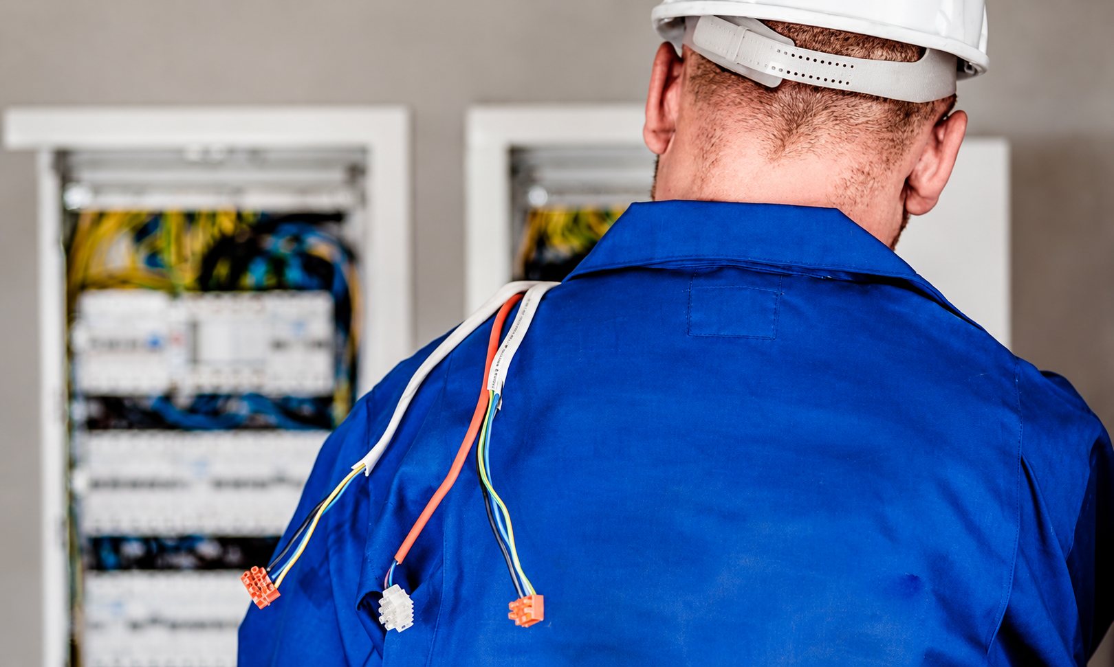 Un électricien en bleu de travail et casque blanc, vu de dos, regarde des armoires électriques ouvertes avec des fils colorés posés sur son épaule.
