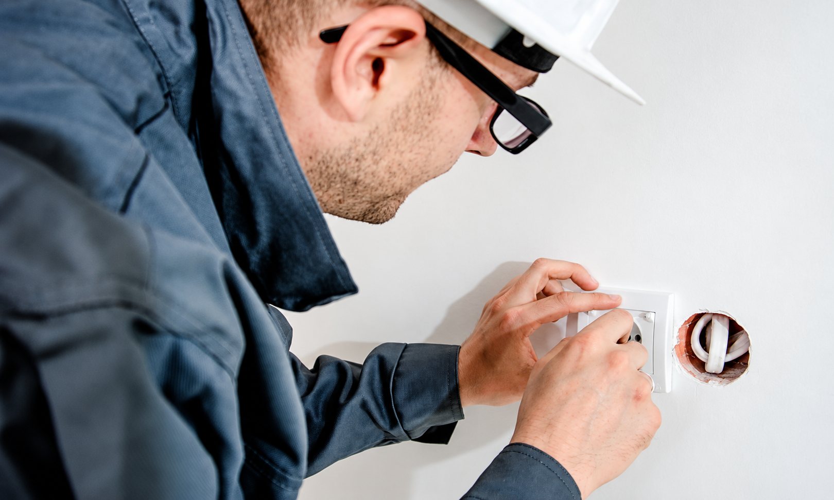 Un électricien portant un casque blanc et des lunettes fixe une prise électrique encastrée dans un mur blanc.