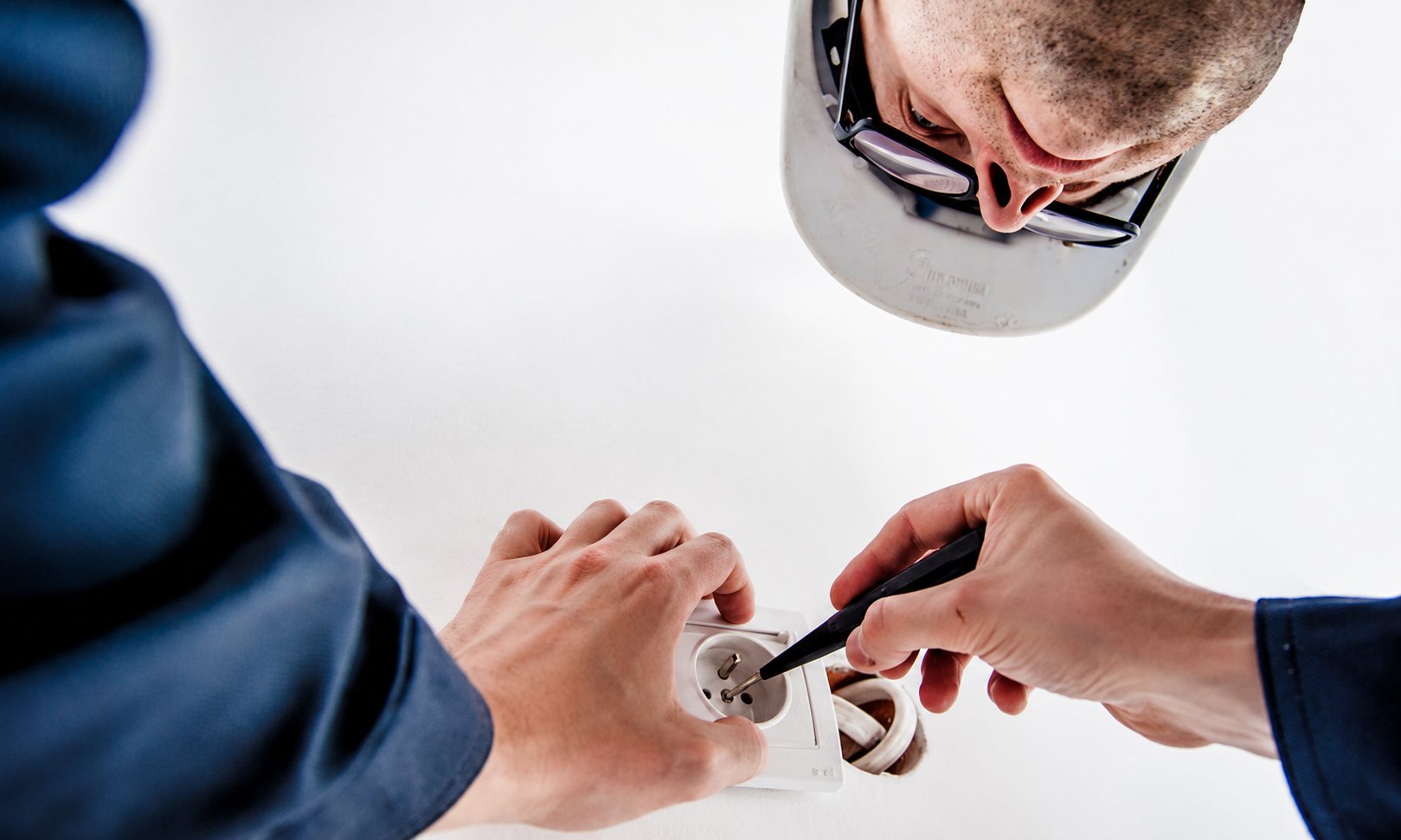 Vu d'en bas, un électricien portant un casque et des lunettes fixe une prise électrique blanche sur un mur avec un tournevis.