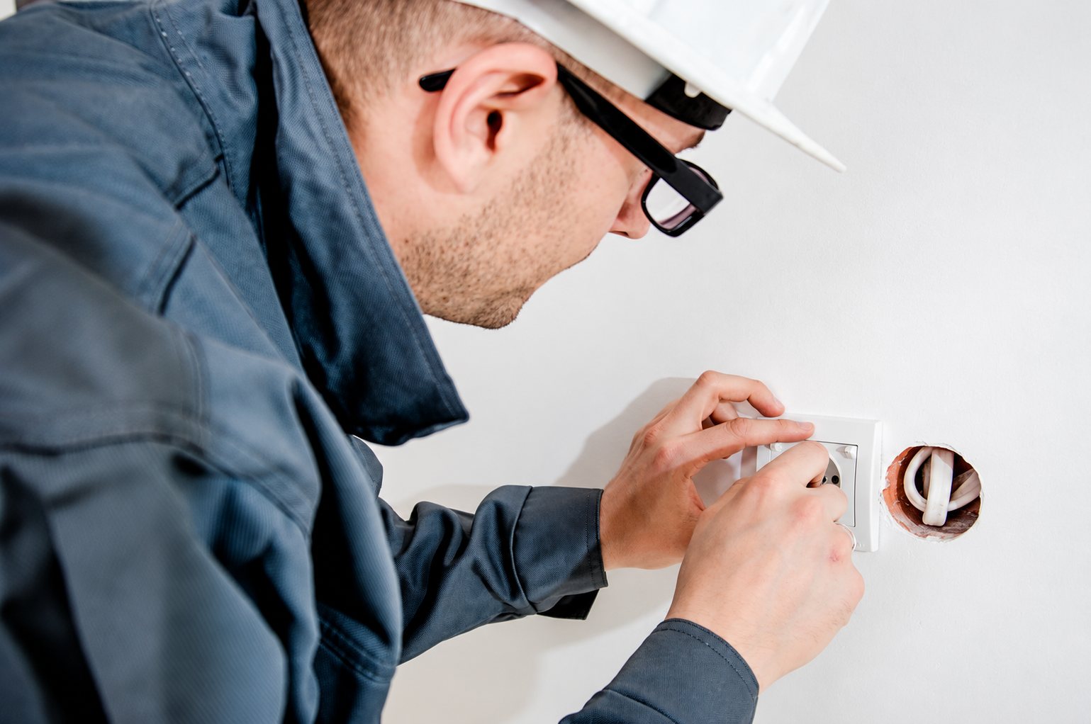 Un électricien portant un casque blanc et des lunettes installe une prise électrique murale.