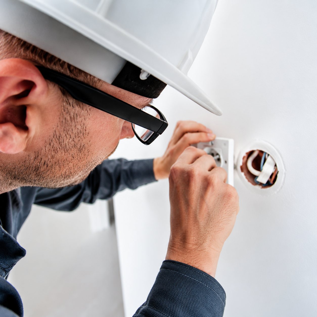 Gros plan d'un ouvrier portant un casque blanc qui intervient avec un outil sur une installation murale.