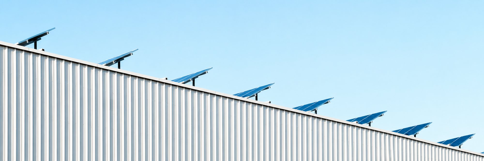 Une rangée de panneaux solaires inclinés installés en haut d'un mur en tôle ondulée blanche sous un ciel bleu clair.